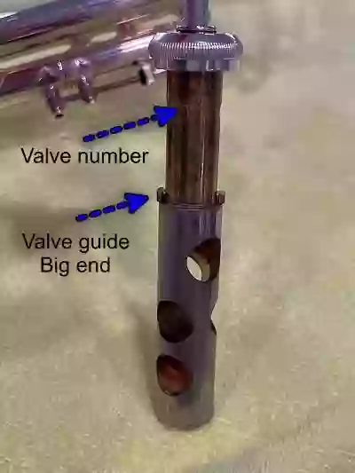 The first piston valve from a Bach trumpet standing on a yellow microfiber cloth with arrows showing the number "1" stamped on the casing and pointing to the Valve guide and the "big end"